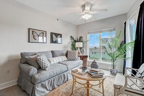 A living room with a grey sofa, two butterfly wall art pieces, a ceiling fan, and a large window with a view of buildings. - Bright, Airy Oceanview Condo on Amelia Island! (Amelia Island)