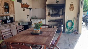 Une table à manger en bois avec des chaises, un coin cuisine doté d’un four en briques et un petit espace extérieur aménagé avec un parasol bleu.