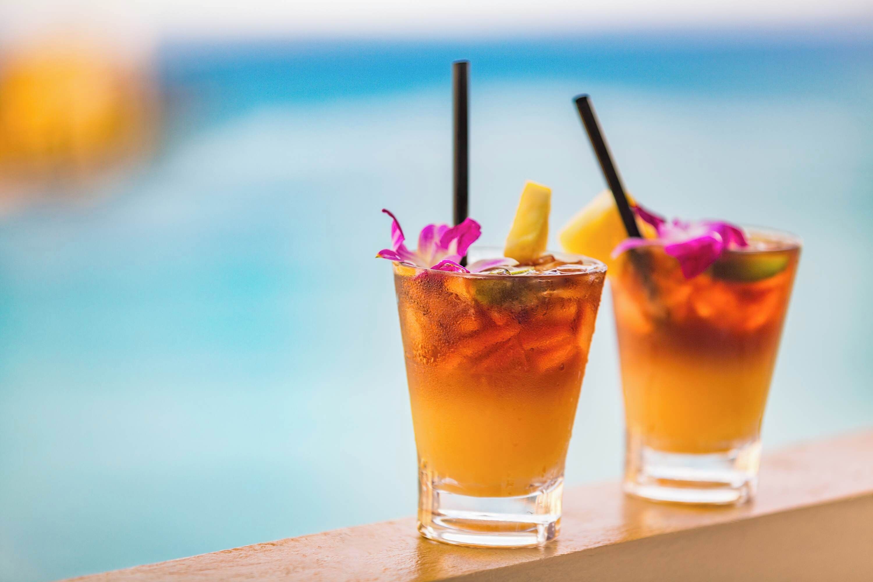 Two cocktails with straws and garnishes, placed on a ledge with a pool in the background.