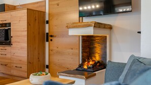 A modern living room with a built-in fireplace, wooden cabinetry, a flat-screen TV, and a dining area with a bowl of fruit. - Comfortable apartment with sauna (Mauterndorf)