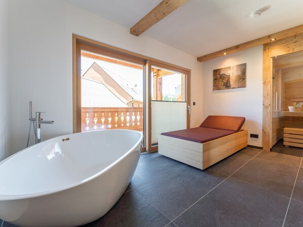 A modern bathroom with a freestanding bathtub, a wooden bench, and a large window offering a view of the outside.
