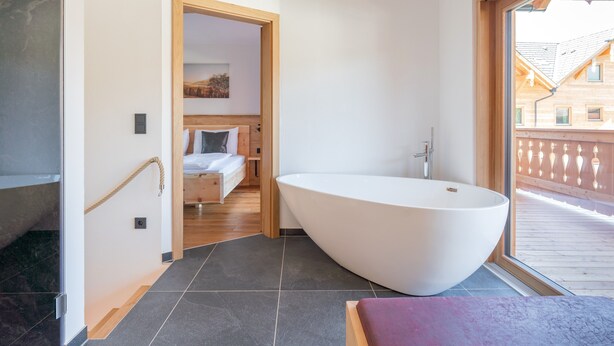 A modern bathroom with a freestanding bathtub, a wooden door leading to a bedroom, and a large window offering a view of the outside. - Exclusive apartment with private sauna (Mauterndorf)