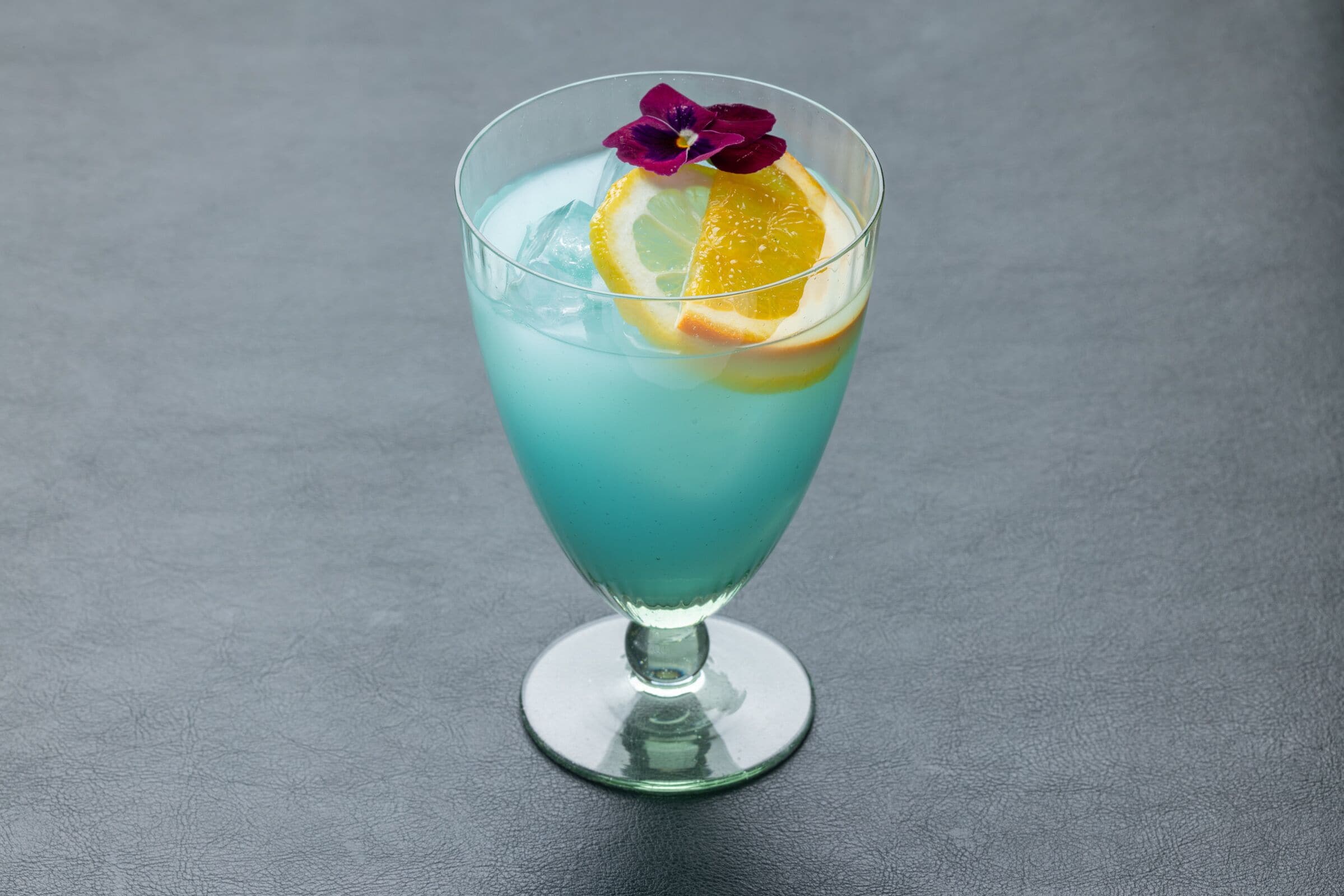 A cocktail glass with a blue liquid, ice, a slice of orange, and a purple flower garnish on a dark surface.