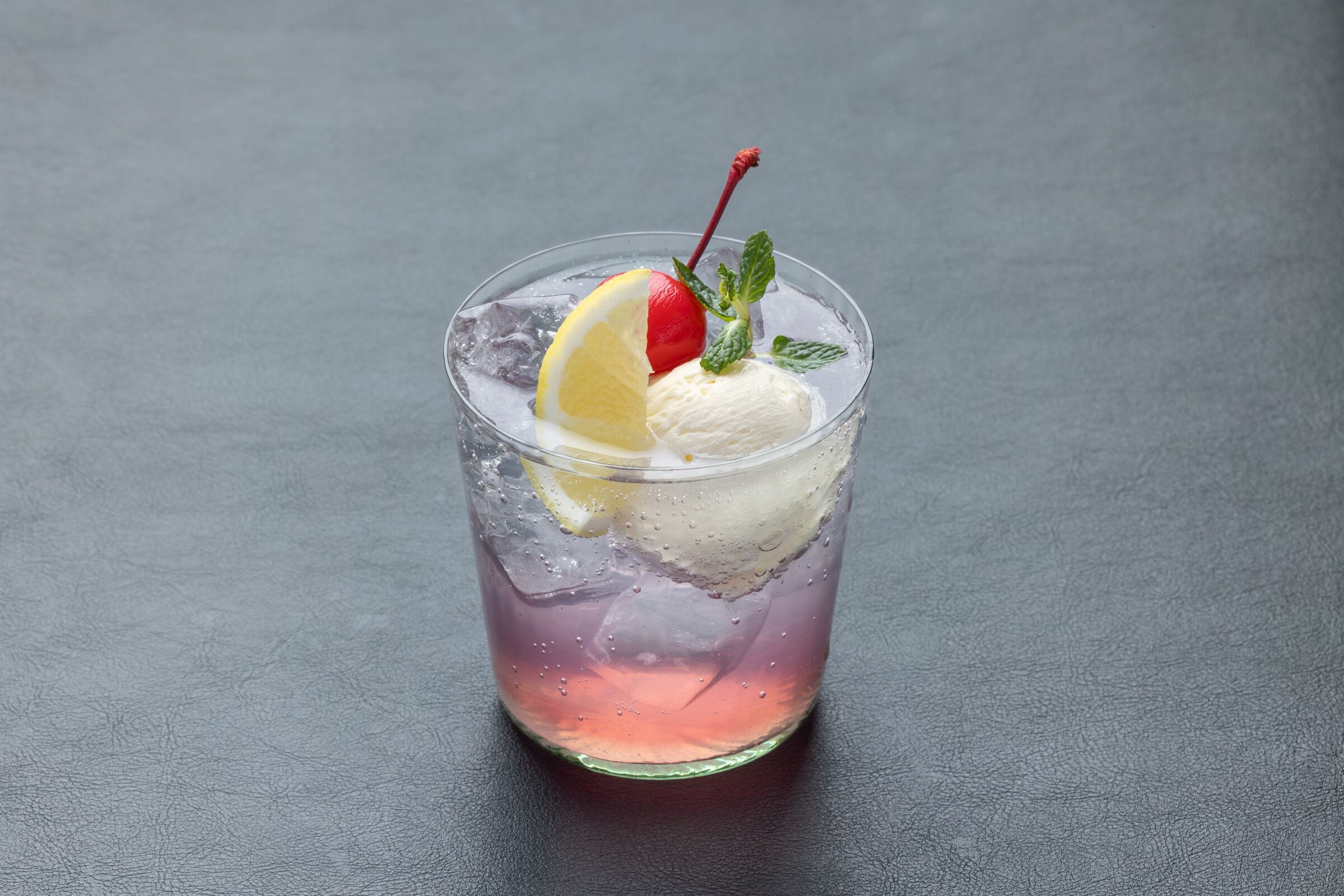 A glass with a purple beverage, a slice of lemon, a cherry, and a scoop of ice cream.