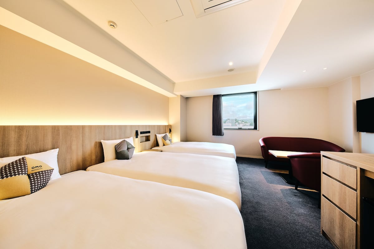 Ett hotellrum med tre sängar, ett skrivbord, en stol och ett stort fönster med utsikt över staden.