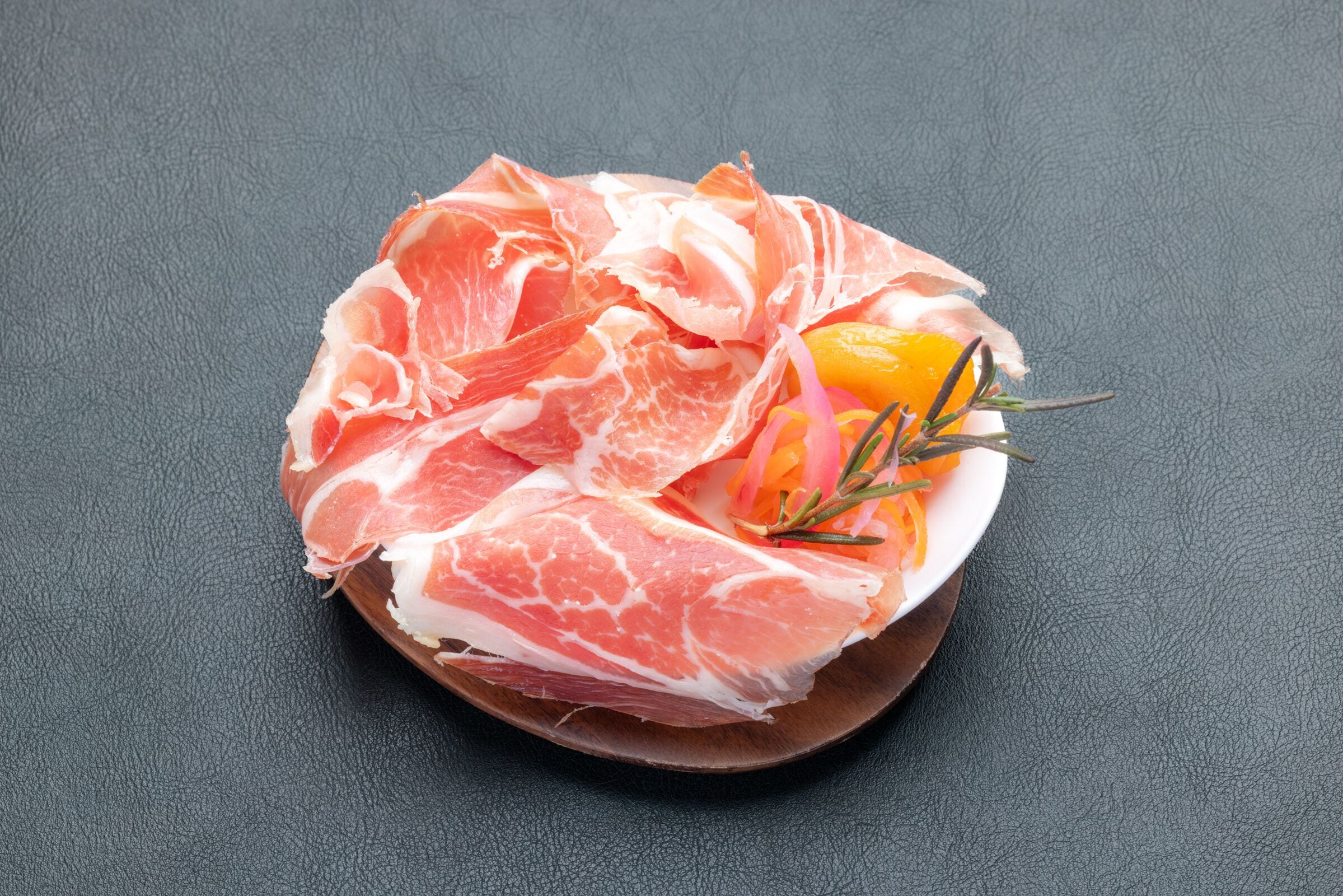 A plate of prosciutto with orange slices and rosemary on a dark surface.