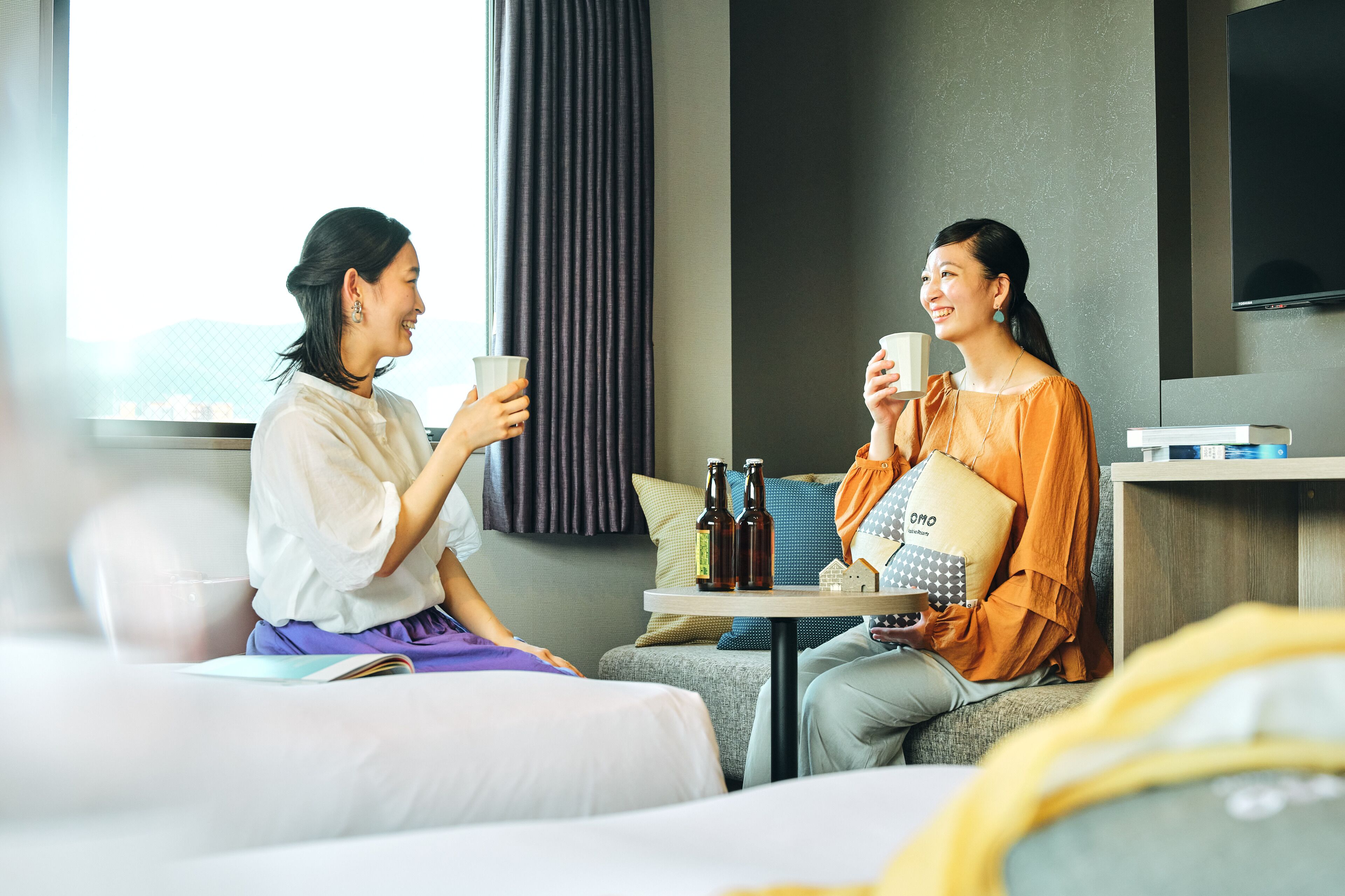 Two people sitting on a sofa in a hotel room, holding cups, with a table between them containing bottles and a pillow.