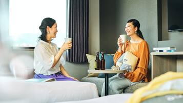 Two people sitting on a sofa in a hotel room, holding cups, with a table between them containing bottles and a pillow.