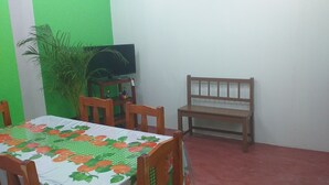Ein Essbereich mit einem Tisch, der mit einer Tischdecke mit Obstmuster bedeckt ist, Holzstühlen, einem Fernseher auf einem kleinen Tisch und einer Topfpflanze.