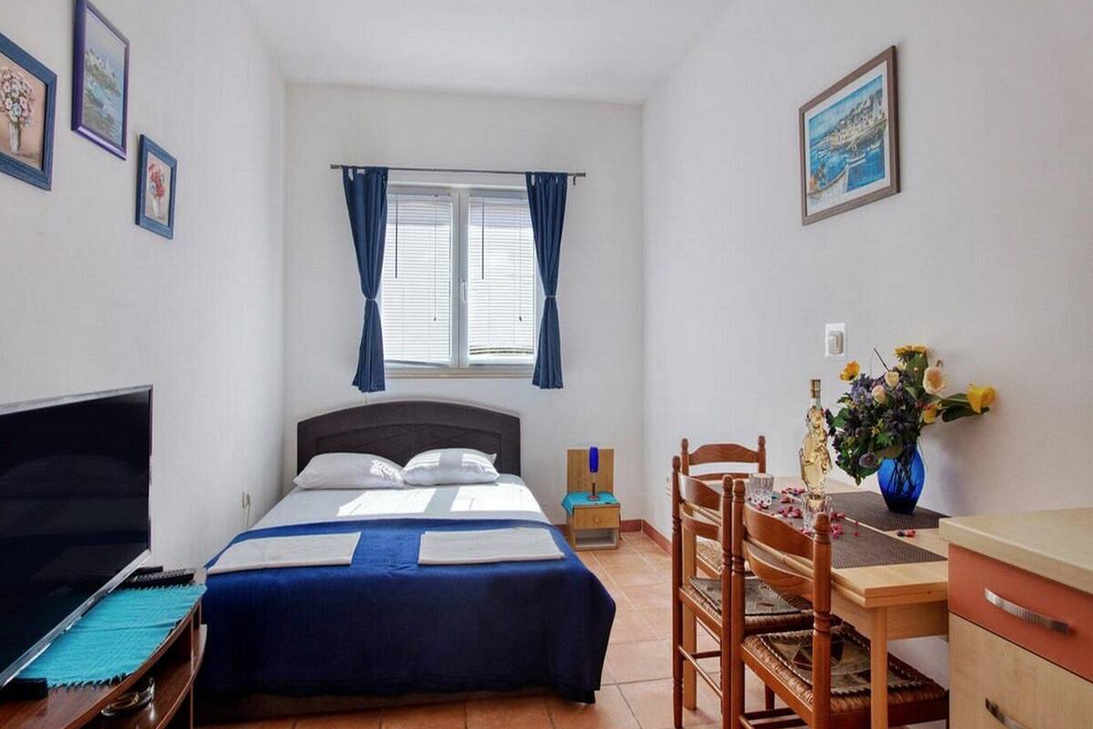 Quarto com cama, escrivaninha, cadeira, televisão e janela com cortinas.