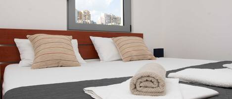 A neatly made bed with a rolled towel, white pillows, and a view of buildings through a window.