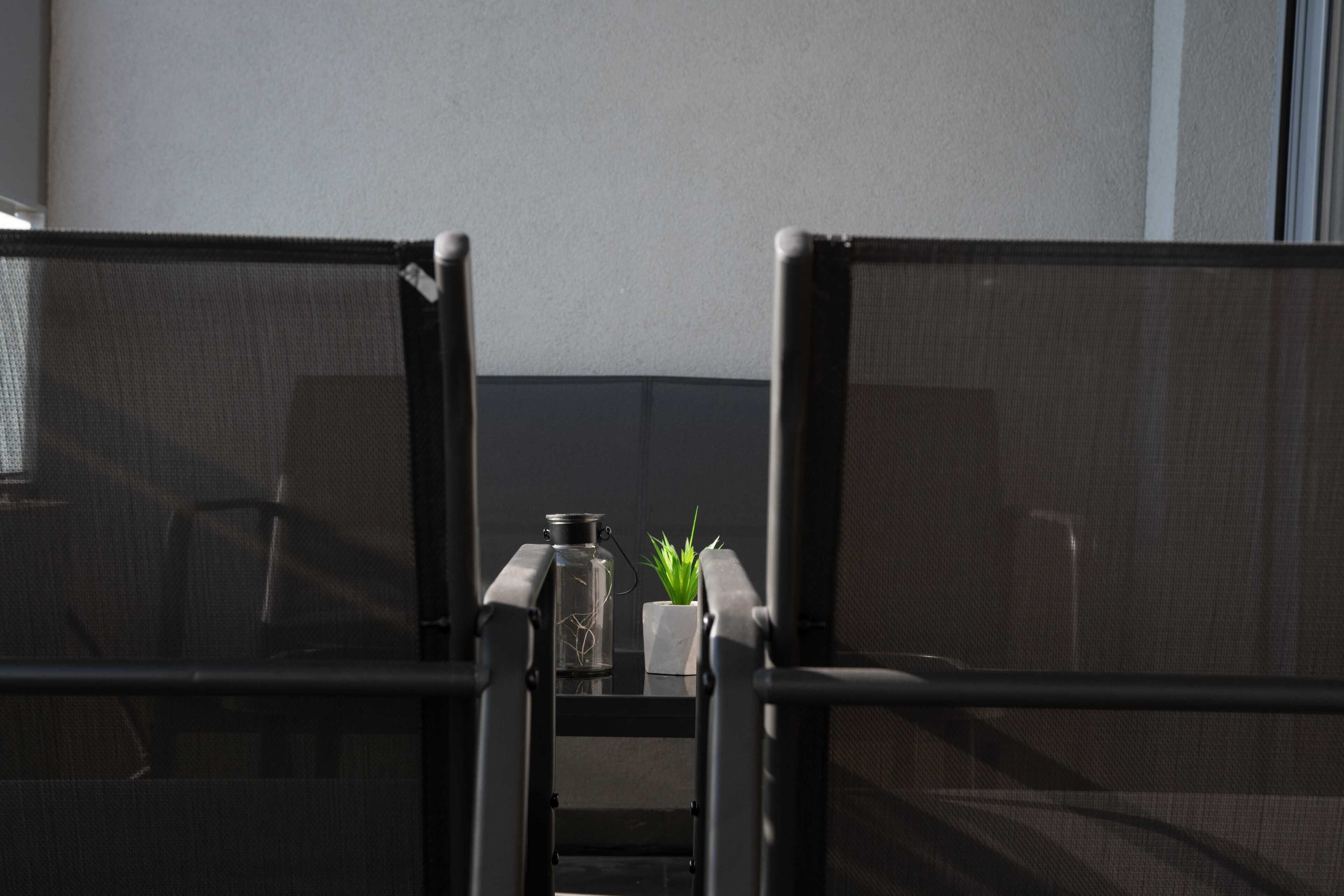 A view through a window with a metal railing, a small table with a glass bottle and a potted plant, and a chair in the background.