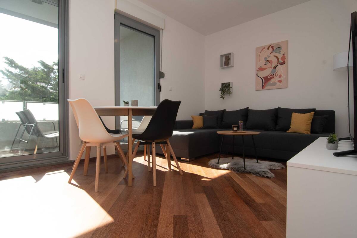 A modern living room with a wooden floor, a black sofa, a white desk, and a dining area with chairs.