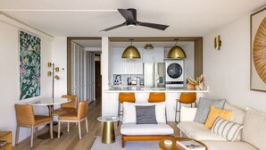 A modern living room with a sofa, coffee table, and chairs. There is a dining area with a table and chairs. The room has a ceiling fan and pendant lights.