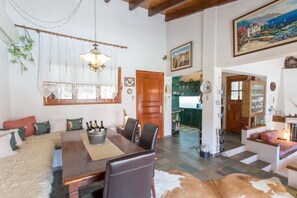 Un vaste salon comprenant une table à manger en bois, des fauteuils en cuir, un canapé agrémenté d’un plaid en fourrure et un tapis en peau de vache. On y trouve également un lustre, un tableau accroché au mur et, au fond, un coin cuisine.