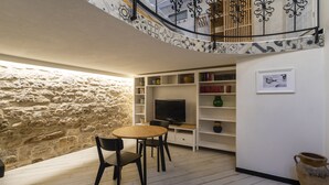 Uma sala de estar acolhedora com parede de pedra, mesa de madeira e cadeiras pretas. Há uma TV em uma prateleira branca e um vaso grande no chão.