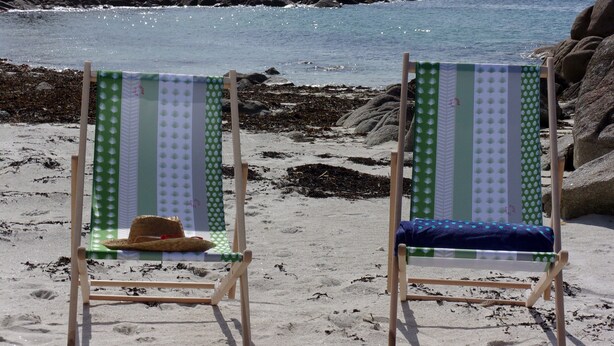 To strandstole med stribet stof, hvoraf den ene har en hat, placeret på en sandstrand med sten og hav i baggrunden.