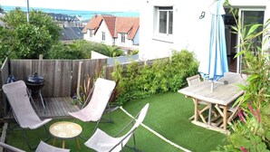 A backyard with a wooden deck, a barbecue grill, two folding chairs, a small round table, and a wooden table with an umbrella. - Sea view, terrace. Sleeps 4. (Fécamp)