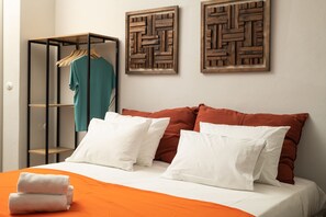 Un lit bien fait, avec une literie blanche et orange, une armoire en bois où sont suspendus des vêtements, et deux œuvres d’art en bois accrochées au mur.