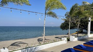 A beachside lounge with blue lounge chairs, a string of lights, and a view of the sea. - Beach Club Lombok by Bale Solah (Senggigi)