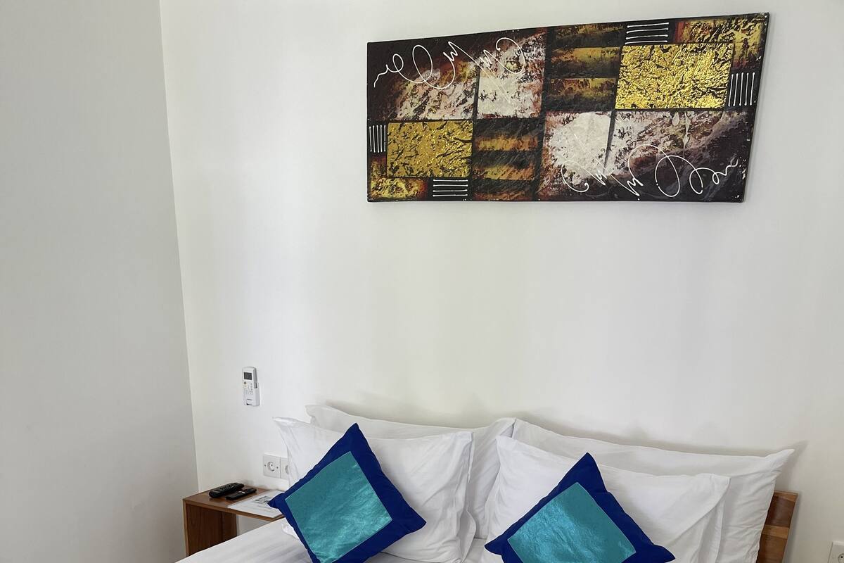 A neatly made bed with white bedding and blue accents, a bedside table with a phone and remote control, and a wall-mounted artwork above the bed.