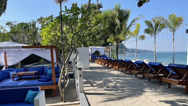 A beachside lounge area with blue cushioned seating, a concrete planter, and a view of the ocean. - Beach Club Lombok by Bale Solah (Senggigi)