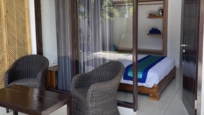 A room with a bed, a wooden chair, a table, and a sliding glass door leading to an outdoor area. - Beach Club Lombok by Bale Solah (Senggigi)