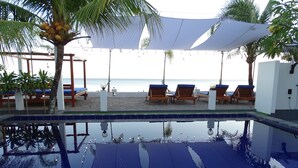 A poolside area with lounge chairs and umbrellas, a palm tree, and a view of the beach. - Beach Club Lombok by Bale Solah (Senggigi)