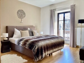 A bedroom with a bed, nightstands, a lamp, a fur throw, a white rug, a window with curtains, and a wall art piece. - Belle Kitz urban villa for 11 with sauna & garden (Kitzbühel)