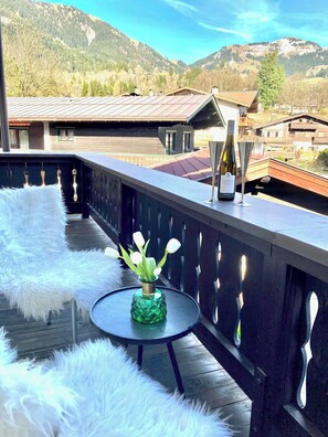 Un balcon avec vue sur les montagnes, une table en verre, un vase rempli de fleurs et une bouteille de vin.
