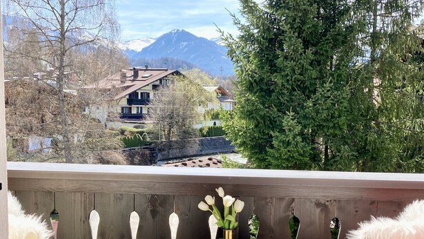 Ein Balkon mit Tisch, auf dem Kuchen und Blumen stehen, ein Sessel mit einem flauschigen Kissen und im Hintergrund eine Aussicht auf Häuser und Berge.