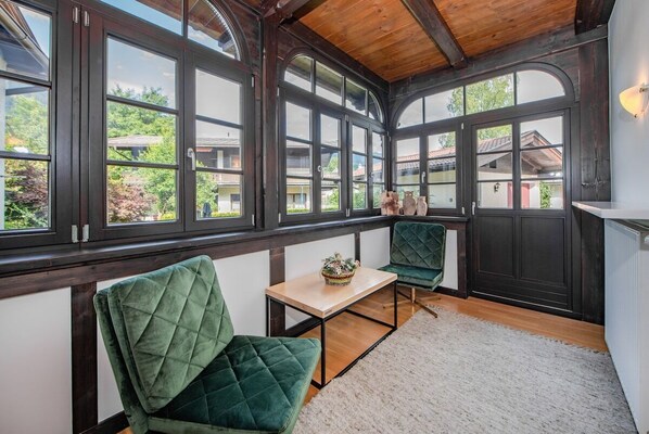 A conservatory with large windows, a wooden ceiling, and green upholstered chairs.