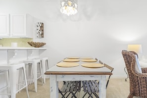 A dining area with a table set for four, white bar stools, a woven chair, and a chandelier.