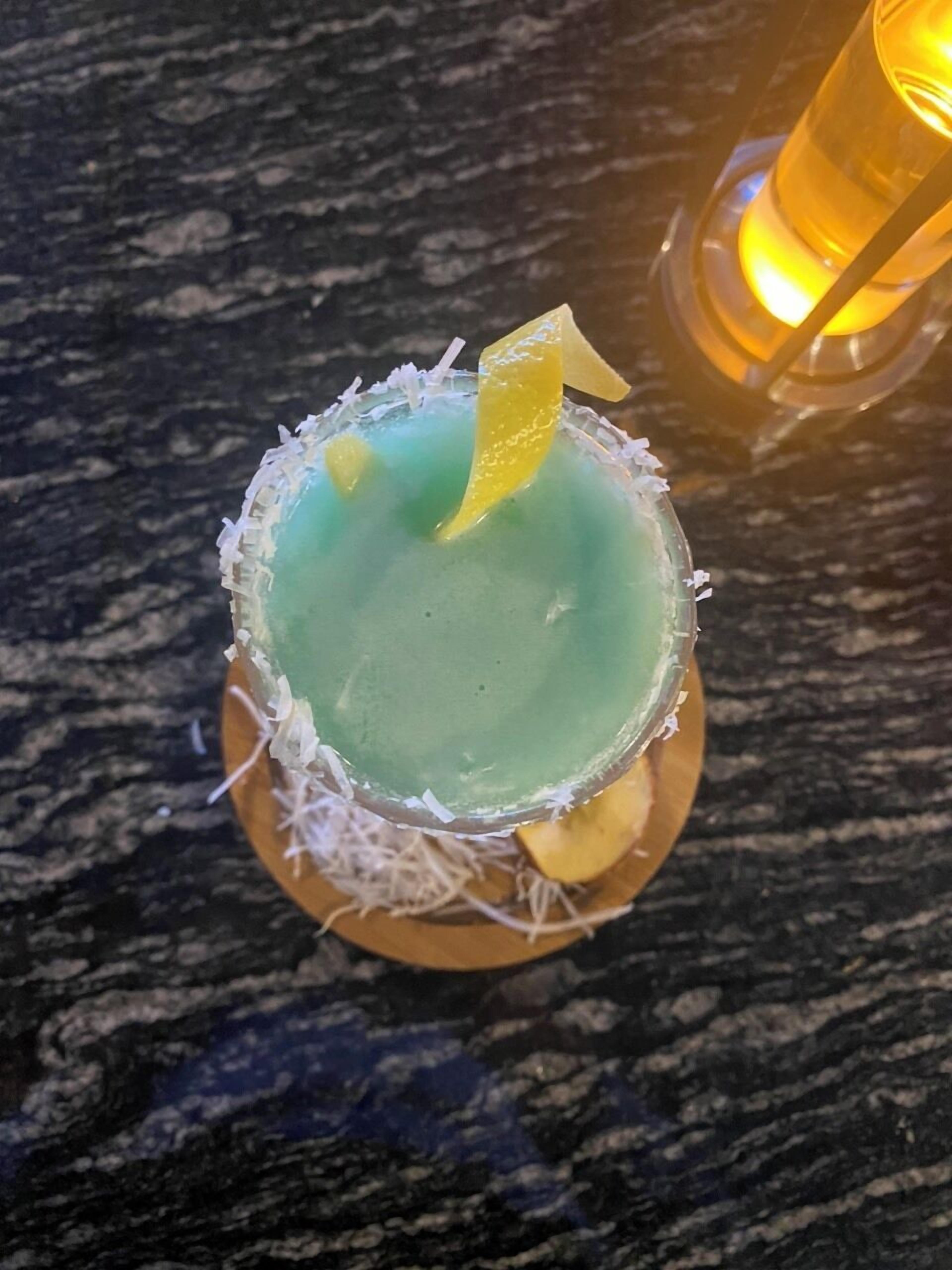 A cocktail in a glass with a salted rim, garnished with a slice of lime and a piece of fruit, on a wooden coaster.
