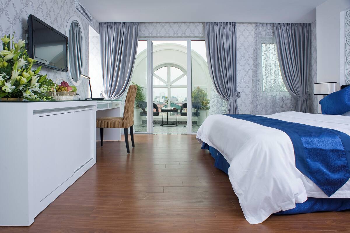 A modern hotel room with a large bed, a desk with a TV, and a balcony with a view.