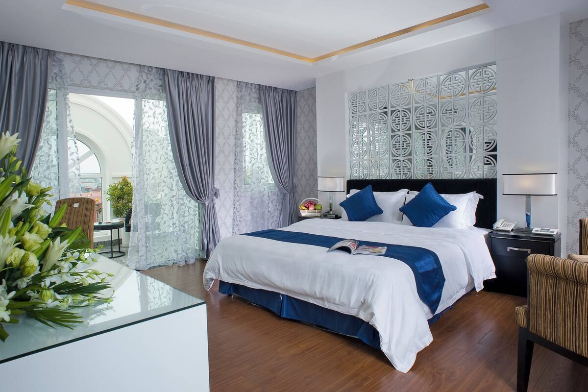 A modern hotel room with a large bed, a glass table with flowers, a nightstand with a lamp, and a decorative wall panel.