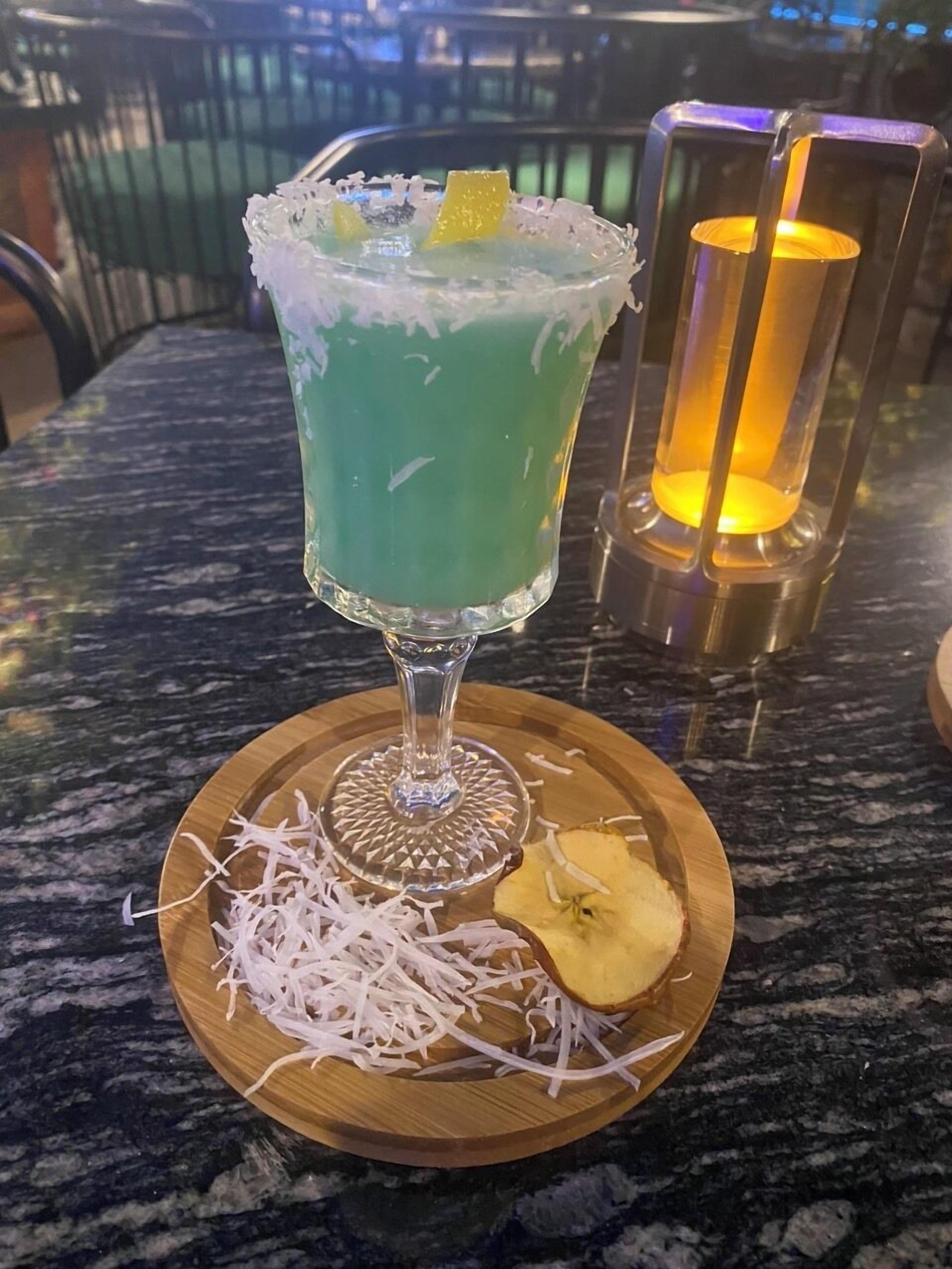 Een cocktail in een glas, met een limoenpartje, versierd met geraspte witte substantie, op een houten onderzetter met een ananaspartje en geraspte witte substantie.
