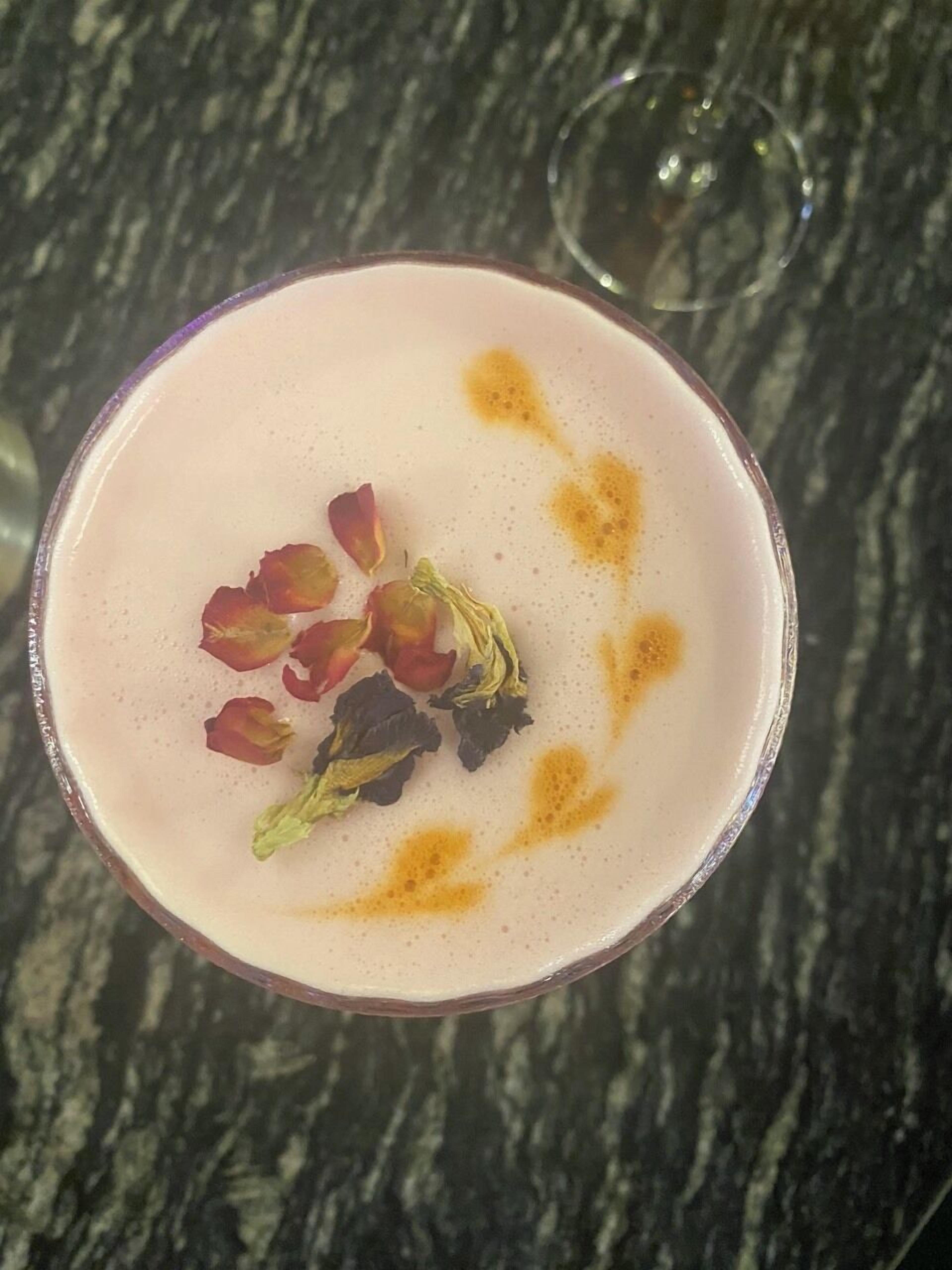 A cocktail with rose petals and a sprig of greenery on a frothy surface.