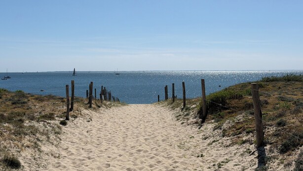 Ein Sandweg am Strand, der zum Meer führt, mit Booten in der Ferne.