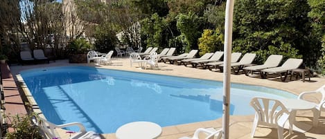 A swimming pool with lounge chairs and tables, surrounded by greenery and buildings.