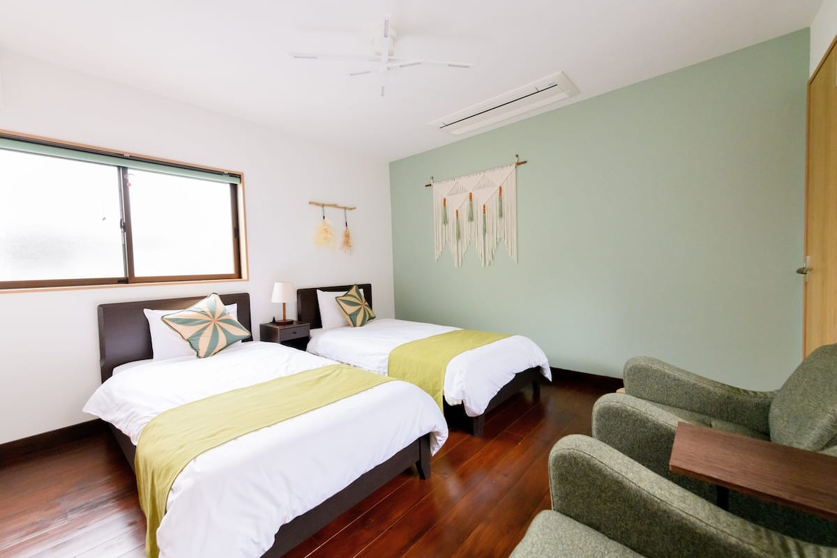 A hotel room with two beds, a green armchair, a wooden floor, a macrame wall hanging, and a large window.