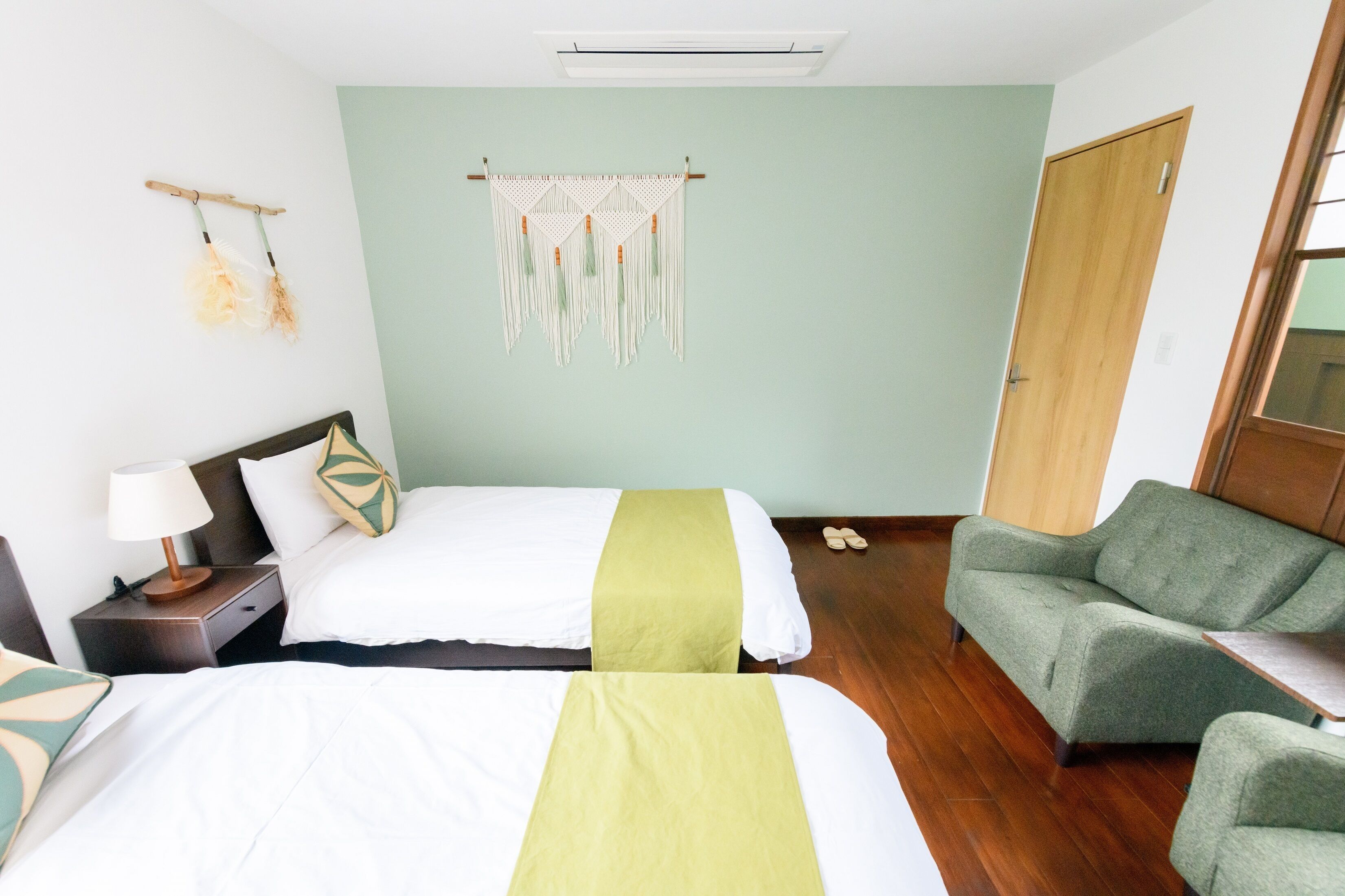 Une chambre d’hôtel avec deux lits, un fauteuil vert, une table de chevet en bois avec une lampe et une décoration murale.
