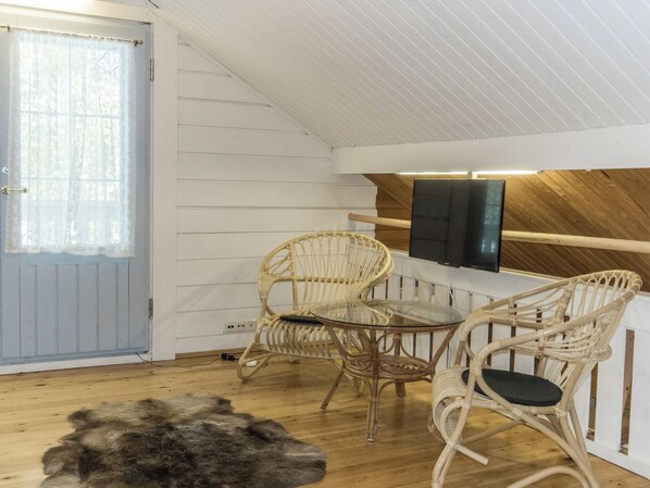 A room with a wooden ceiling, a television mounted on the wall, a glass table, wicker chairs, and a fur rug on the floor.