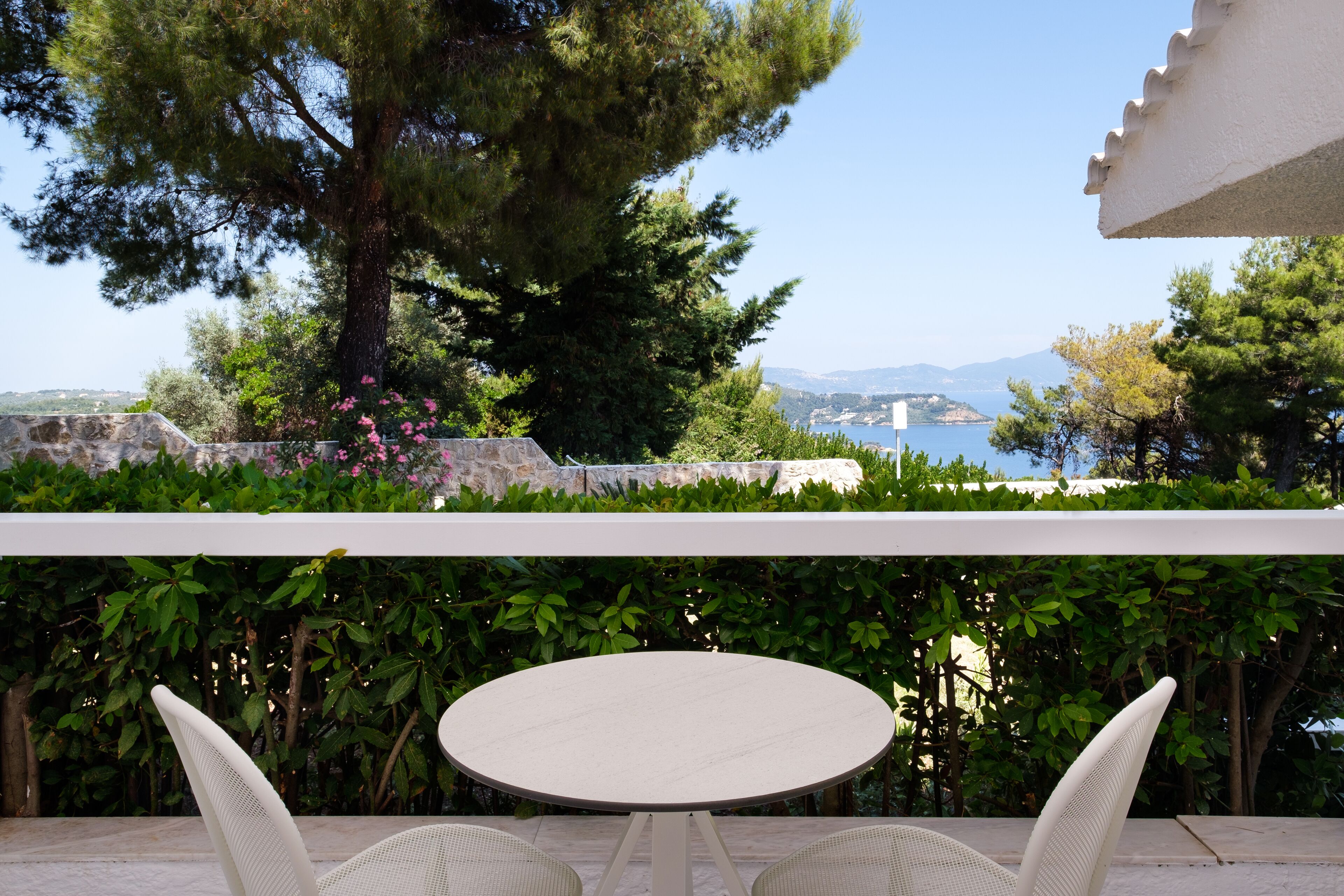 An outdoor patio with a round table and white wicker chairs, overlooking a scenic view of a body of water and mountains.
