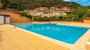 Une piscine à l’eau d’un bleu cristallin, entourée d’une terrasse carrelée. Au fond, on aperçoit des bâtiments dotés de balcons et un paysage vallonné.