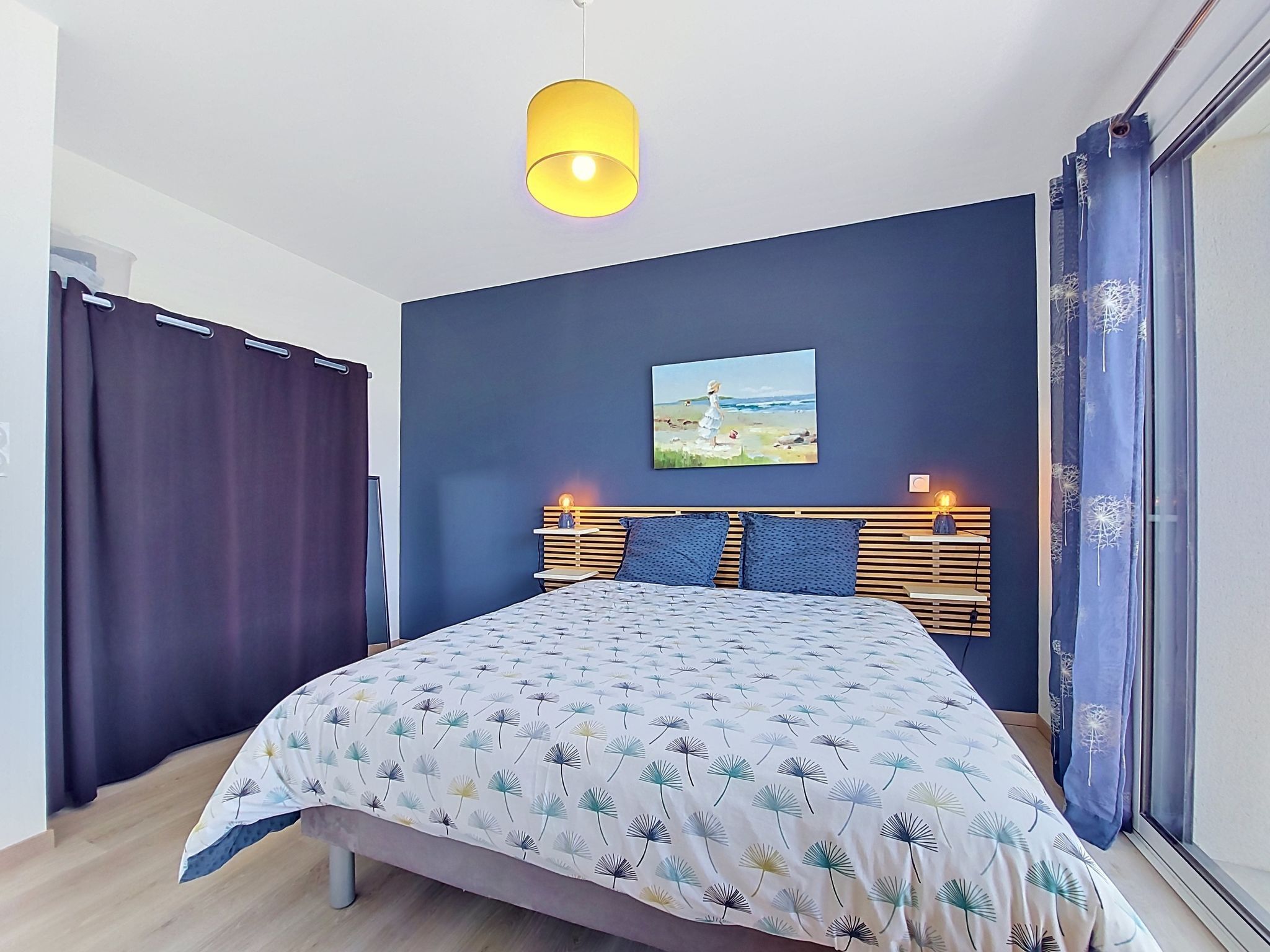 A bedroom with a bed, a painting, a bedside table, and a hanging light fixture.