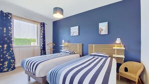 Une chambre avec deux lits, une fenêtre avec des rideaux, une chaise et des œuvres d’art accrochées au mur.