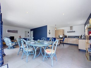 Une cuisine moderne avec des meubles en bois, un coin repas avec une table ronde et des chaises bleues, et un salon avec un canapé et un téléviseur.