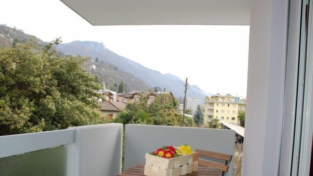 A balcony with a wooden table and chairs, a small table with fruit, and a view of a mountain and buildings. - Simona Fontile 22 by Interhome (Minusio)