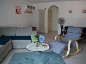 En stue med en sofa, en lænestol, et rundt bord med en vase blomster og en ventilator.